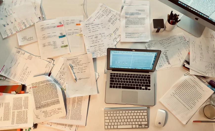 Ein SChreibtisch von oben, darauf steht ein Laptop und drumherum liegen beschriebene A4-Zettel, Bücher und Ausdrucke noch und nöcher
