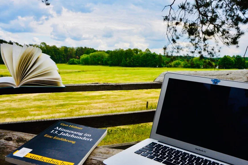 Im Vordergrund ein Laptop, daneben ein Buch "Minnesang im 13. Jahrhundert". Auf einem Holzgeländer ein weiteres Buch aufgeschlagen. Blick in die Landschaft über ein gemähtes Feld, dahinter Bäume. Am Himmel blaue Quellwolken
