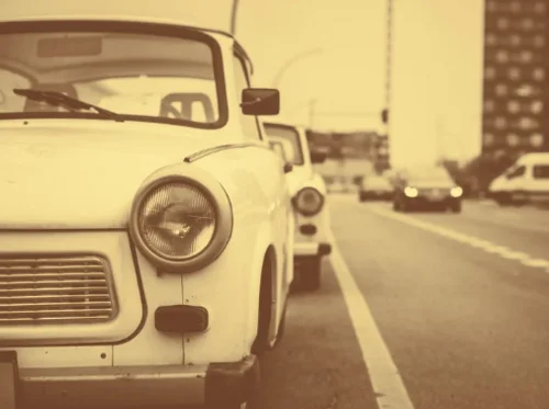 Zu sehen ist in Sepia-Farben ein Trabant 601, der am Seitenrand einer Straße steht. Hinter ihm ist die rechte Seite eines weiteren Trabis zu sehen.