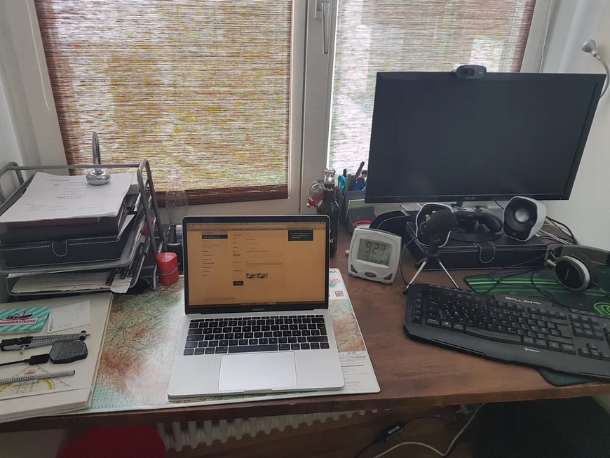 Schreibtisch vor einem Fenster. in der Mitte ein aufeklappter Laptop, rechts danaben ein zweiterBildschirm und eine Tastatur, links eine Dokumentenablage