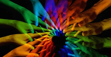 Button zur Unterseite Weitere Interessenvertretungen: Das Bild zeigt eine dynamische Darstellung mehrerer Hände, die sich kreisförmig ausstrecken und sich fast in der Mitte berühren. Die Hände leuchten in verschiedenen Regenbogenfarben (Blau, Grün, Gelb, Orange, Rot) und überlagern sich teilweise, vor einem dunklen Hintergrund mit einem schwarzen Punkt in der Mitte.