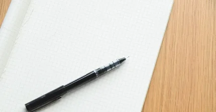 Auf einem Holztisch liegen ein leeres weißes Blatt und ein schwarzer Gelschreiber.