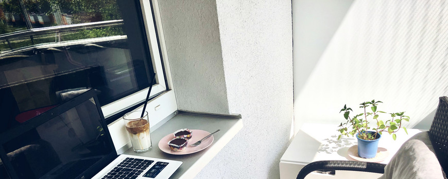 Ein Laptop steht auf einem Fensterbrett, außen auf einem balkon, vor dem FEnsterbrett steht eine Couch. daneben liegen zwei kleine Kuchen und ein Glas Kakao steht auch da