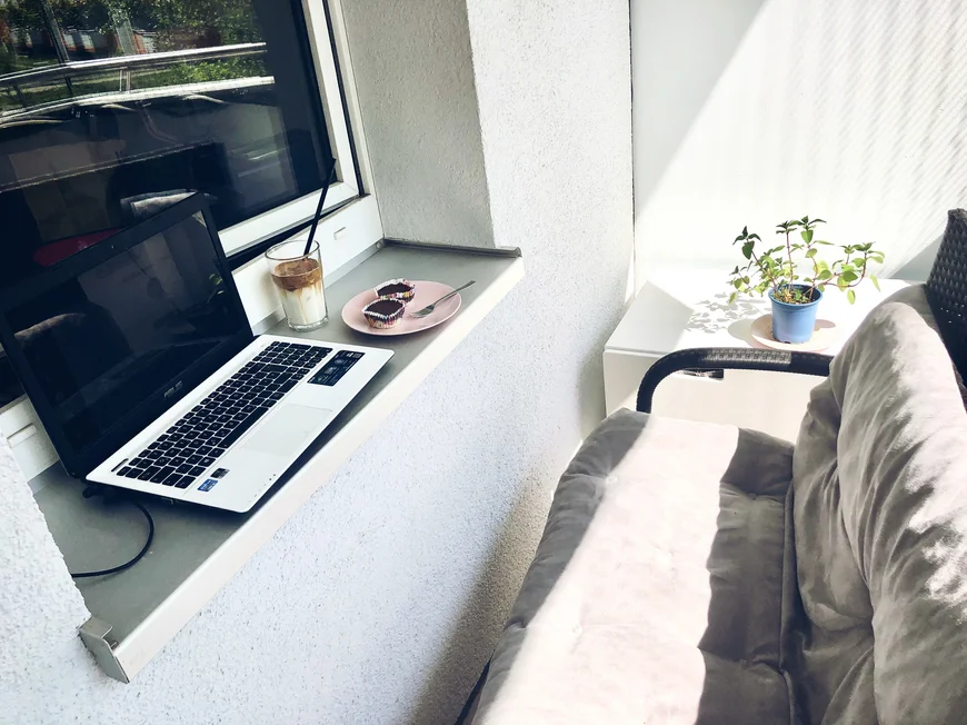 Ein Laptop steht auf einem Fensterbrett, außen auf einem balkon, vor dem FEnsterbrett steht eine Couch. daneben liegen zwei kleine Kuchen und ein Glas Kakao steht auch da