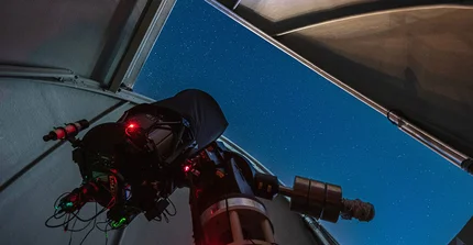 Blick in den Sternenhimmel aus der Kuppel einer Sternwarte. Im Vordergrund ist ein großes Teleskop zu erkennen.