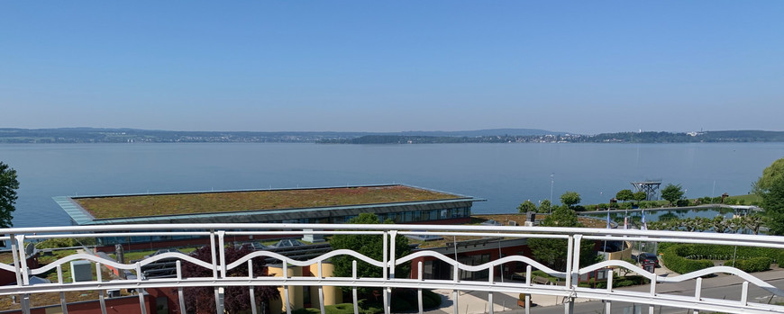 von einem Balkon aus forografiert. Blick über einen großen SEe, man sieht das gegenüberliegende Ufer. Im Vordergrund ein Balkongeländer und auf einem Tisch steht ein Laptio