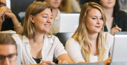Zwei Studentinnen sitzen mit einem Laptop in einem Hörsaal und lächeln. Um sie herum sitzen weitere Studierende.