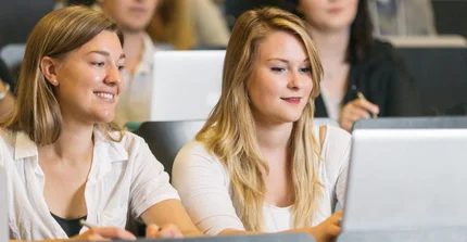 Zwei Studentinnen sitzen mit einem Laptop in einem Hörsaal und lächeln. Um sie herum sitzen weitere Studierende.
