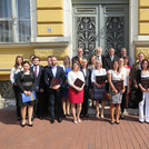 Eine Gruppe mit ca. 20 Absolventinnen und Absolventen in sommerlich feierlicher Kleidung sowie Professorinnen und Professoren steht im Freien vor dem Eingangsportal des gelben Gebäudes der Juristischen Fakultät der Universität Szeged.