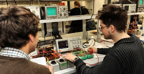 Zwei Personen arbeiten an einem physikalischen Experiment in einem Labor. Der Versuchsaufbau besteht aus vielen Kabeln und einigen elektronischen Geräten.