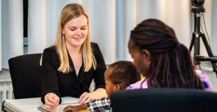 Eine junge Frau macht ein kognitionswissenschaftliches Experiment mit einem Kind, das bei seiner Mutter auf dem Schoß sitzt