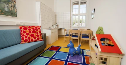 The picture shows the parent-child room: a bright, cheerfully furnished room with a blue sofa and red cushions, a colorful carpet, toys such as a blue bouncing horse and a wooden dollhouse. At the back there is a small dining area with a table and chairs and a mini kitchen. A large window lets in plenty of daylight.