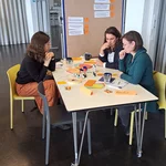 Verschiedene Menschen sitzen an Tischen im Rahmen eines Workshopsettings und haben viele verschiedene Materialien auf dem Tisch.