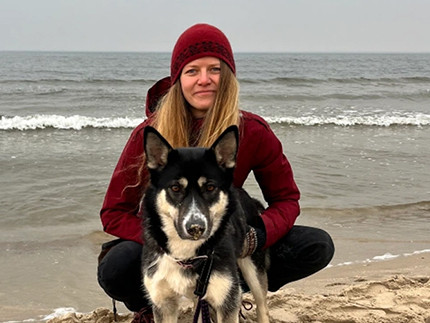 Leonna Szangolies sits squatting on the beach. The sea begins just behind her. In front of her is a dog who is also looking towards the camera. The sky is overcast and the weather is cool.