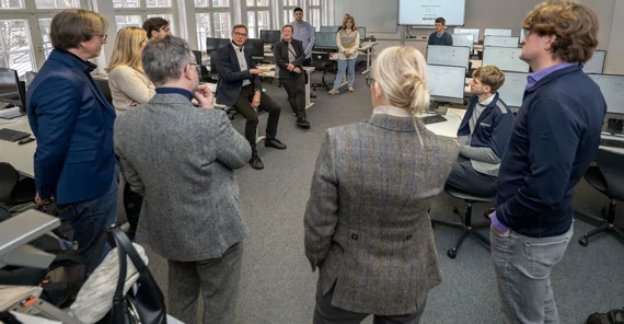 Mehrere Personen stehen in einem Computerpool im Kreis und hören einer Person aus dem Kreis zu
