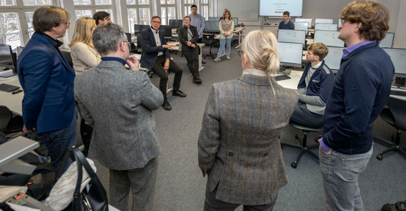 Mehrere Personen stehen in einem Computerpool im Kreis und hören einer Person aus dem Kreis zu