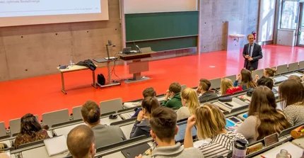 Ein Mann steht in einem Hörsaal vor vielen Studierenden und hält eine Vorlesung