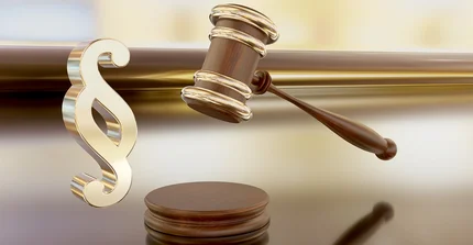 Denken Sie an eine Darstellung, die das Konzept von Recht und Gerechtigkeit symbolisiert. Im Vordergrund sehen Sie ein großes, glänzendes Zeichen – das ist das Paragraphen-Symbol, das aussieht wie ein doppeltes "S". Es ist aus goldglänzendem Metall gefertigt und wirkt sehr solide und wichtig. Daneben, leicht erhöht und schräg liegend, ist ein Richthammer zu sehen, auch "Gavel" genannt. Er hat einen runden Kopf und einen Griff, beides aus dunklem, poliertem Holz, und ist mit glänzenden, goldenen Bändern verziert, die ihn umschließen. Unter dem Hammer, direkt auf dem Boden, liegt eine kleine, runde Holzscheibe, auf die der Hammer normalerweise schlagen würde. Der gesamte Hintergrund ist weich und unscharf, in warmen Gold- und Brauntönen gehalten, die das glänzende Gold des Paragraphen-Symbols und des Hammers hervorheben. Das Bild vermittelt einen ernsten und offiziellen Eindruck, der mit juristischen Angelegenheiten oder Entscheidungen assoziiert wird.