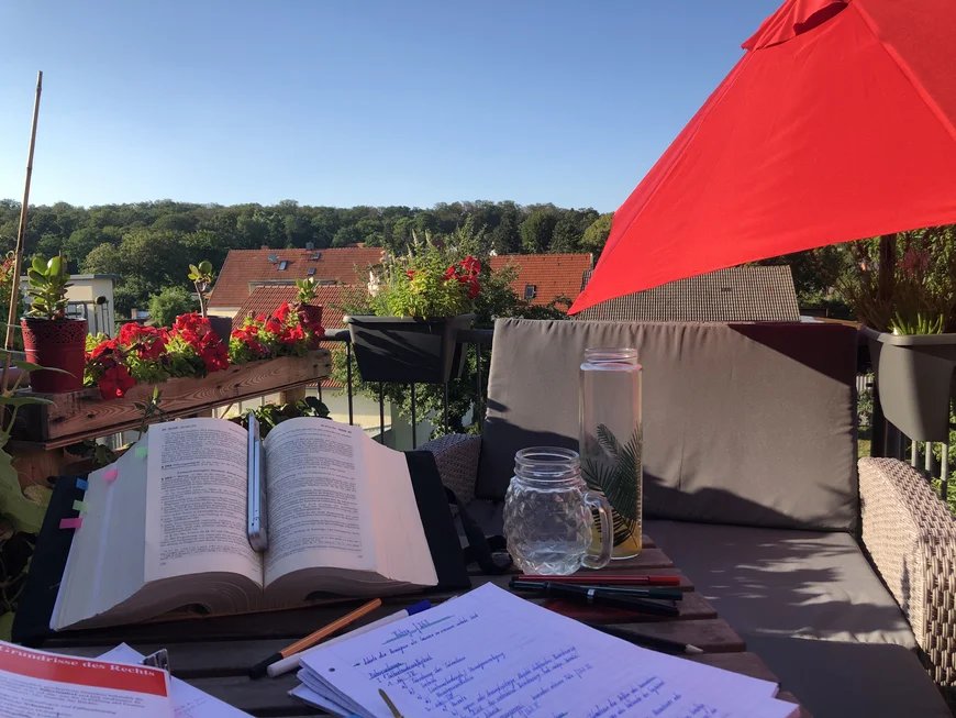 Im vordergrund ein mit Büchern und Schreibsachen vollgepackter Schreibtisch auf einem Balkon, der Blick geht über ein paar Dächer hinweg in eine waldige Landschaft. Der Himmel ist blau und sonnug