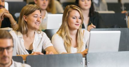 Zwei Studentinnen sitzen mit einem Laptop in einem Hörsaal und lächeln. Um sie herum sitzen weitere Studierende.