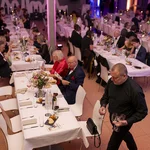 Tische mit weißen Tischdecken, an denen mehrere Personen sitzen.
