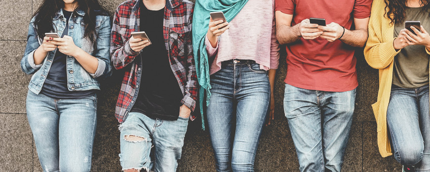 Fünf junge Menschen stehen an eine Hauswand gelehnt nebeneinander und halten Smartphones in der Hand. Sie sind ab den Knien bis zum Hals zu sehen.