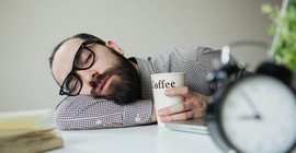 Mann mit schwarzer Hornbrille legt seinen Kopf auf dem Tisch ab. In der Hand hält er einen Kaffeebecher. Im Vordergrund befindet sich ein Wecker. Foto: Fotolia/katie martynova.