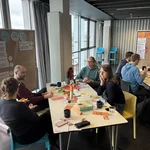 Verschiedene Menschen sitzen an Tischen im Rahmen eines Workshopsettings und haben viele verschiedene Materialien auf dem Tisch.
