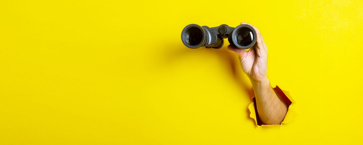 Yellow surface with an arm sticking through a hole. The corresponding hand holds binoculars up to the yellow surface, making it look as if there could be a pair of eyes behind it. - 