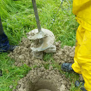 Foto: meadow with borehole filled with muddy shallow groundwater. people with rain gear and auger standing next to it | Foto: Cosmic Sense consortium