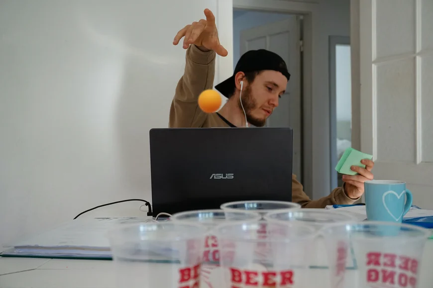 Im Vordergrund stehen einige leer Platikbierbecher. Weiter hinten sitzt ein Student vor seinem Laptop. Sein Blick richtet sich auf Karteikarten, die er in der linken hand hält. mit der rechten Hand wirft er lässig einen Tischtennisball in Richtung der Becher