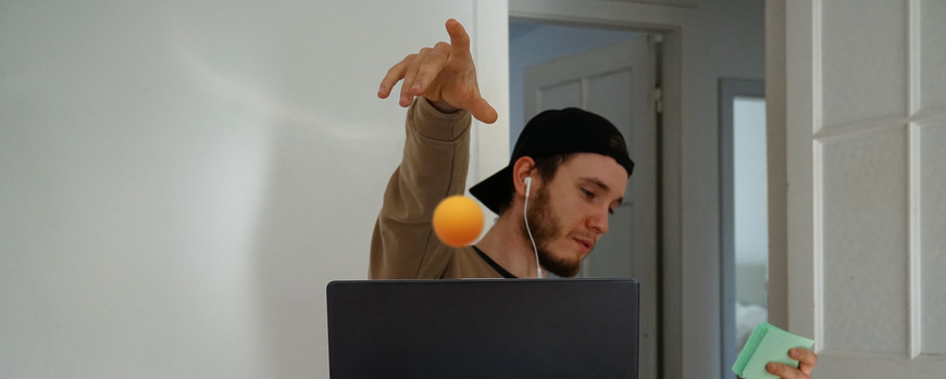 Im Vordergrund stehen einige leer Platikbierbecher. Weiter hinten sitzt ein Student vor seinem Laptop. Sein Blick richtet sich auf Karteikarten, die er in der linken hand hält. mit der rechten Hand wirft er lässig einen Tischtennisball in Richtung der Becher