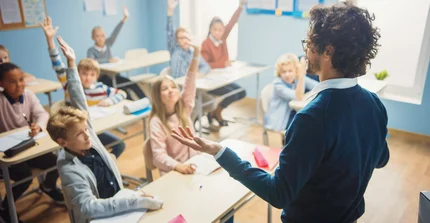 Lehrer steht vor einer Klasse und fragt die Schüler etwas. Diese melden sich begeistert.