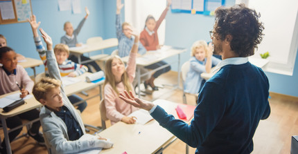 Lehrer steht vor einer Klasse und fragt die Schüler etwas. Diese melden sich begeistert.
