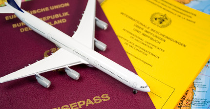 Dokumente mit Reisepass und Impfausweis liegen unter einem kleinen Modellflugzeug. Im Hintergrund schimmert eine Landkarte hervor