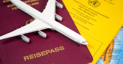 Dokumente mit Reisepass und Impfausweis liegen unter einem kleinen Modellflugzeug. Im Hintergrund schimmert eine Landkarte hervor