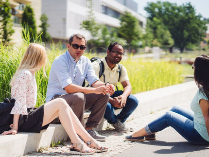 Studierende sitzen auf dem Campus Golm auf einer Bordsteinkante. Hinter ihnen ist eine Wiese zu sehen.