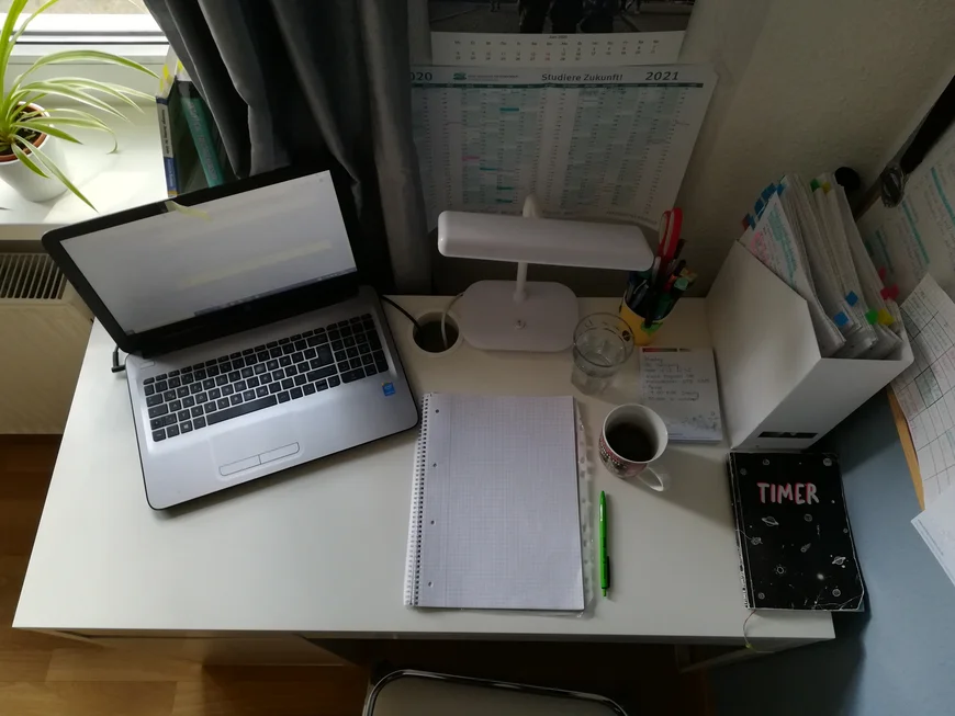 Schreibtisch von oben. Laptio, Lampe, Unterlagen. An der Wand ein Jahreskalender und auf dem schreibtisch ein Büchlein mit der Aufschrift "Timer"