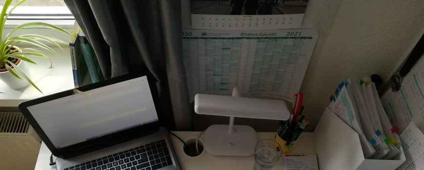 Schreibtisch von oben. Laptio, Lampe, Unterlagen. An der Wand ein Jahreskalender und auf dem schreibtisch ein Büchlein mit der Aufschrift "Timer"