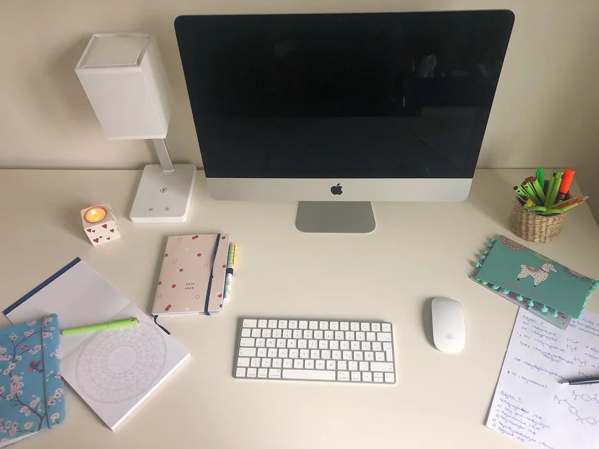 Blick auf einen weißen Schreibtisch. Es gibt einenBildschirm, rechts und links der Tastatur sind Schreibutensilien angeordnet