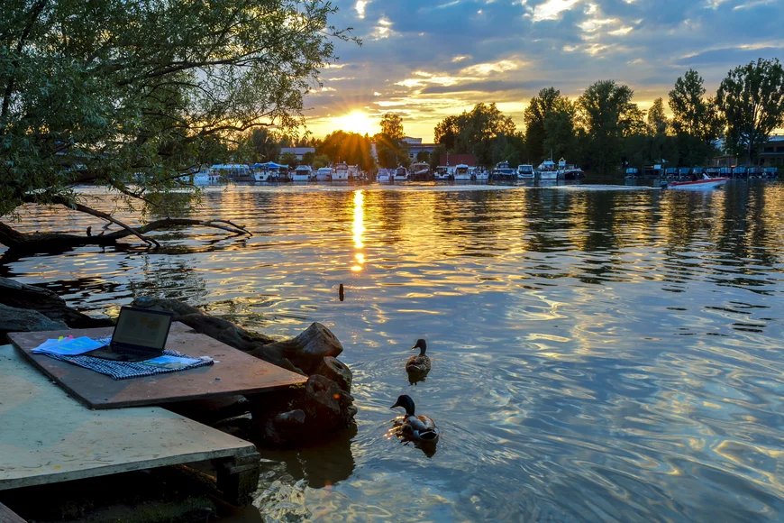 Auf einem provisorischen Steg steht ein Laptop, nah am Ufer schwimmen 2 Enten. DIe untergehende Sonne spiegelt auf der Wasseroberfläche. Auf dem gegenüberliegenden Ufer sind Boote und Bäume zu sehen. Die Sonne bricht durch die WOlken