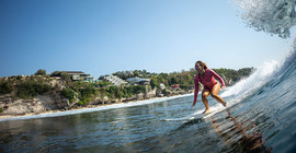 Dr. Anna von Rebay steht auf einem Surfbrett und surft im Meer auf einer Welle.