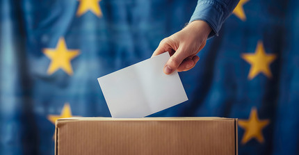 Brief wird in eine Wahlurne gesteckt, dahinter eine Europa-Flagge