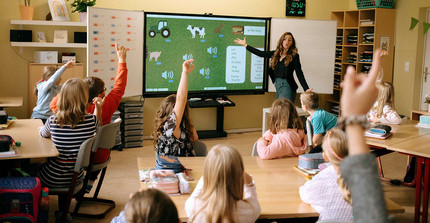 Eine Lehrerin steht vor einer Klasse und erklärt. Viele Kinder melden sich.