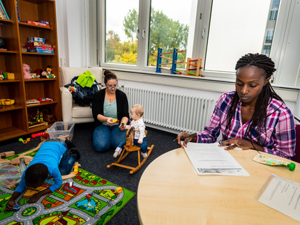 eine Frau spielt mit Kleinkind in einem Zimmer, ein anderes Kind spielt daneben und eine weitere Frau sitzt an einem Tisch und macht Notizen