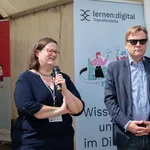 Zwei Personen vor einem Zelt, die linke Person spricht in ein Mikrofon.