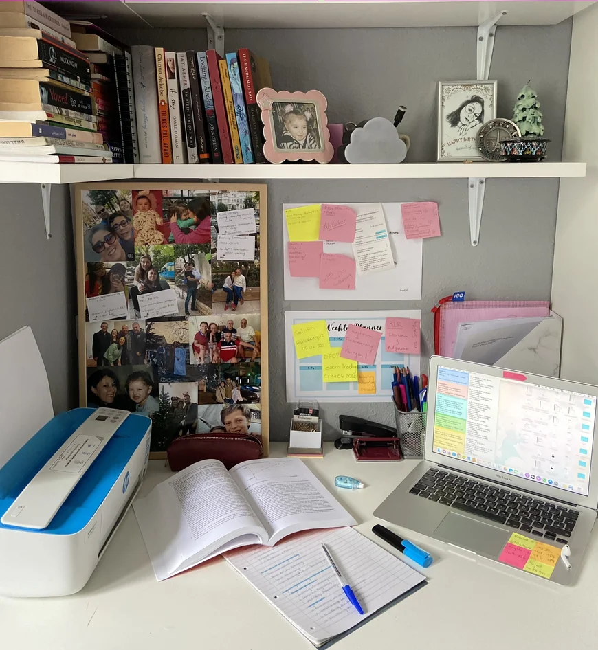 Tisch mit Drucker, Pinnwand mit Fotos von FAmilie und Freunden, post Its, Bücherregal darüber, Bilderrahmen mit Fotos, Schreibteug, Buch, Laptop