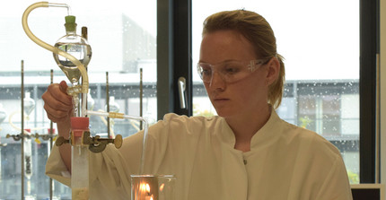 Studierende mit Schutzbrille und Laborkittel in der Hand einen Glaskolben