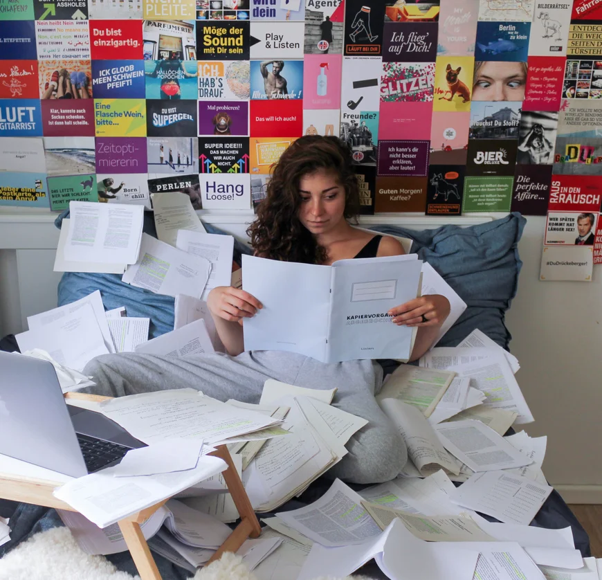 Auf einem Bett, fast begraben unter ausgedruckten Texten und mit einem Laptio vor sich, sitzt eine Studentin und liest. sie sieht nich glücklich aus