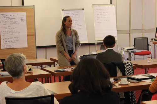 Das Bild zeigt eine Frau, die in einem Seminarraum vor einer Gruppe sitzt und spricht. Sie steht vor Flipcharts mit handschriftlichen Notizen. Mehrere Personen sitzen an Tischen und hören zu. Auf den Tischen liegen Unterlagen, Notizbücher und Getränke.